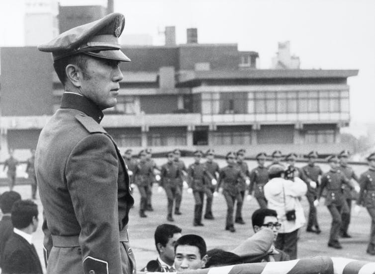 En uniforme militaire, Mishima observe les marcheurs.